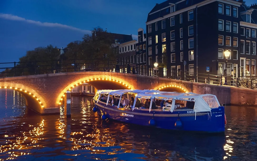 Amsterdam evening canal cruise with passengers enjoying wine and cheese under illuminated bridge.