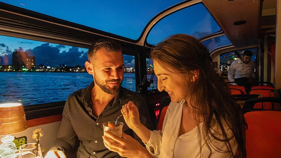 Couple enjoying dessert on an Amsterdam dinner cruise with city skyline in the background.