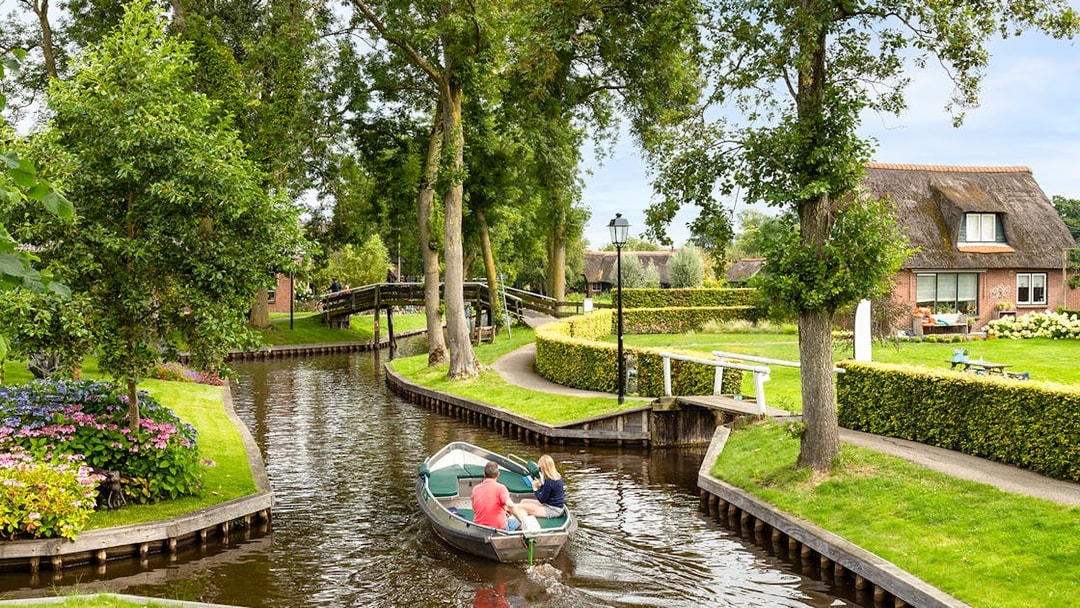 Small electric boat on a canal in Giethoorn, passing under a wooden bridge with thatched-roof houses nearby.
