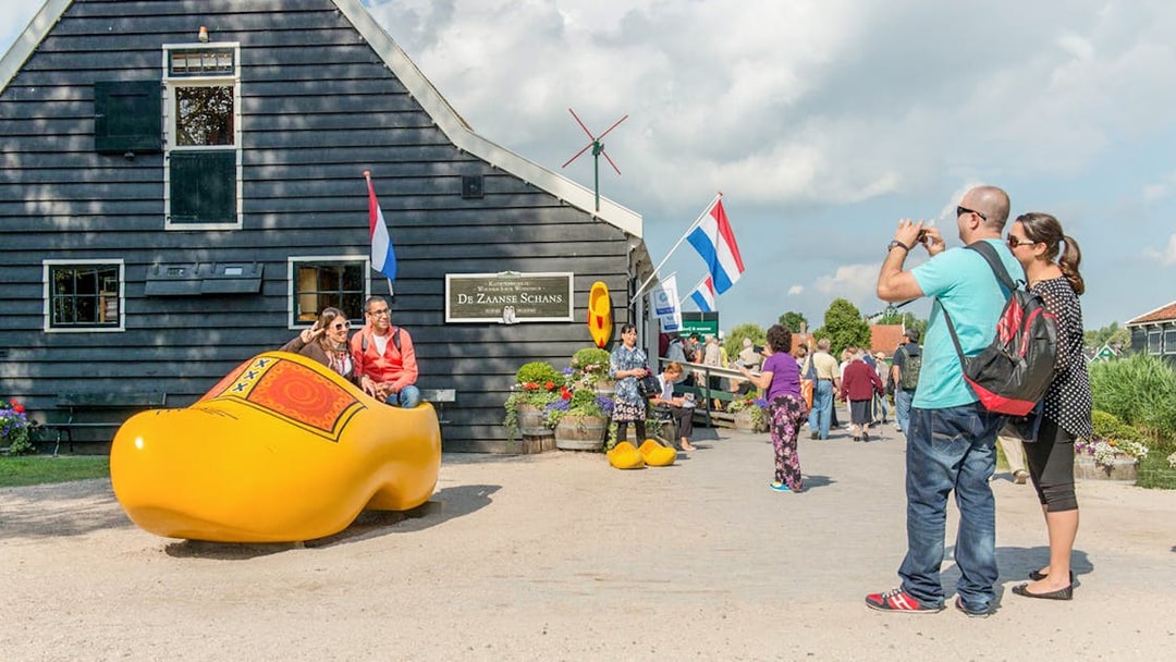 Visitors at Zaanse Schans posing in a giant wooden shoe with Dutch flags and windmill in the background.