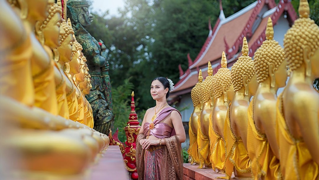 Woman in traditional attire walking between golden Buddha statues at an ancient city temple.