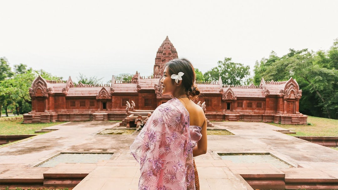 Woman in traditional attire at Ancient City, Thailand, with red temple in background.