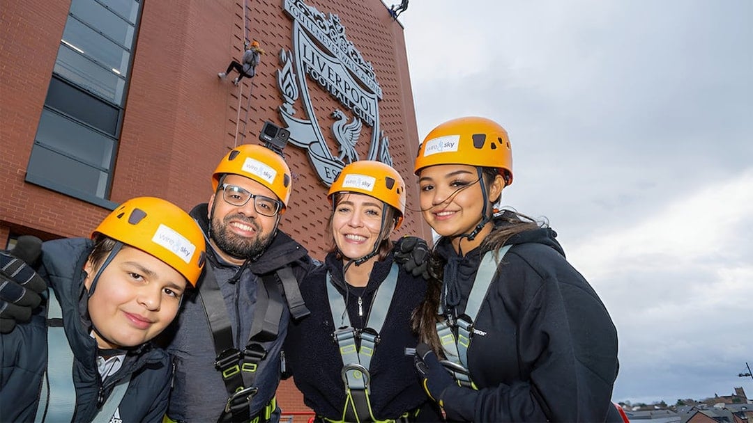 Group preparing for abseil at Liverpool FC's Anfield Stadium with club emblem in background.