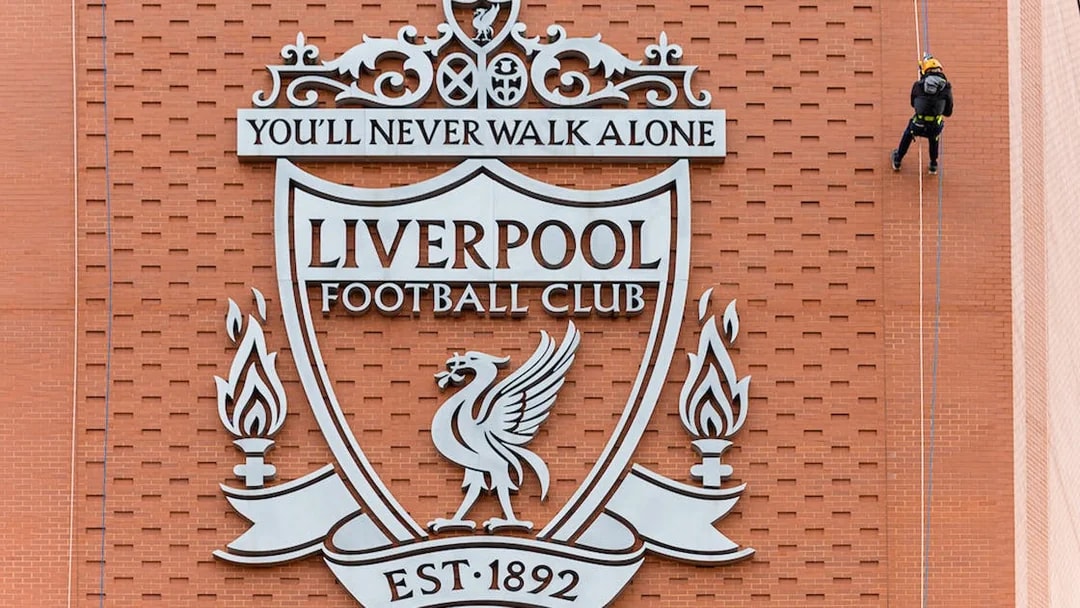 Abseiler descending Anfield Stadium wall beside Liverpool FC crest.