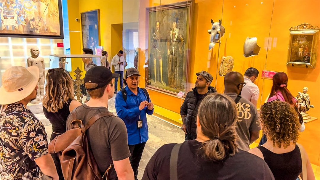 Tour group listening to a guide in the Anthropology Museum, Mexico, with artifacts and paintings displayed.