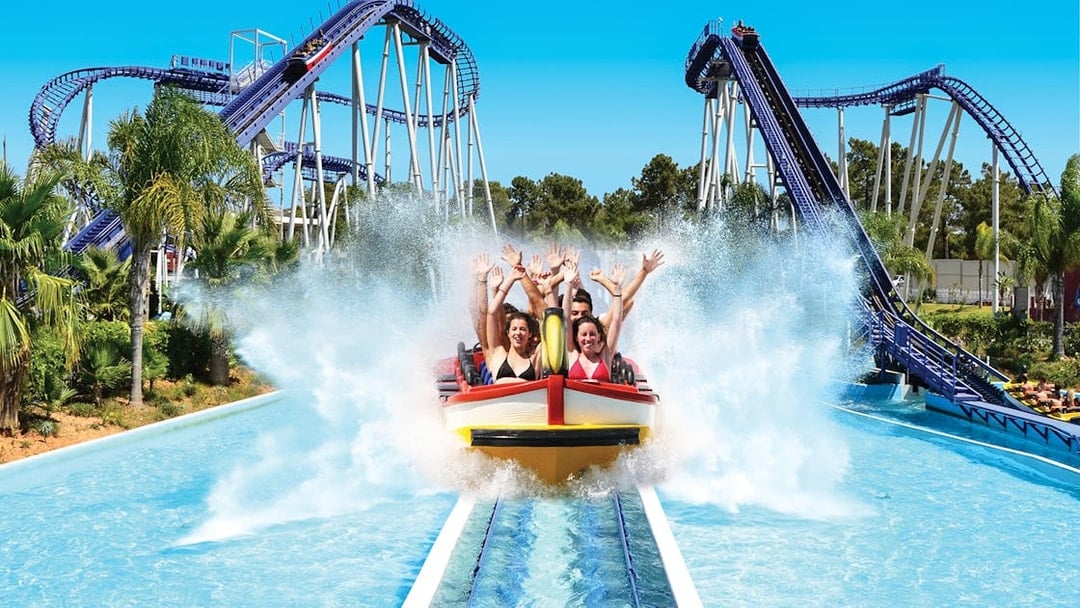 Visitors enjoying a water ride at Aquashow Water Park with roller coasters in the background.