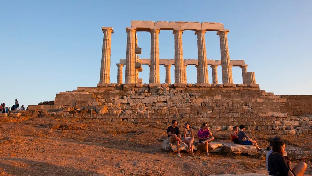 Temple of Poseidon at sunset with visitors seated on ancient stones.