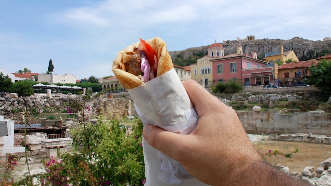 Hand holding a gyro with Athens ruins and colorful buildings in the background.
