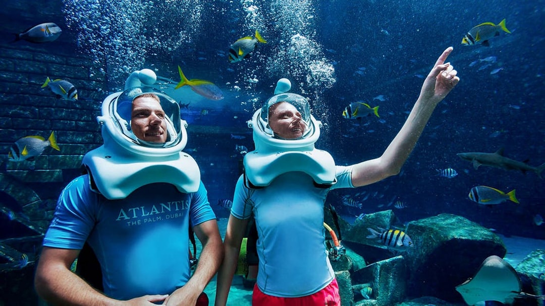 Tourists scuba diving with helmets at Atlantis, surrounded by fish and marine life.