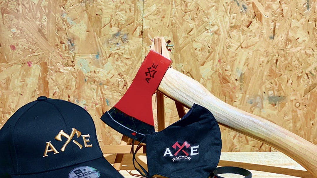 Axe Factor merchandise including a cap, mask, and axe on a wooden background.