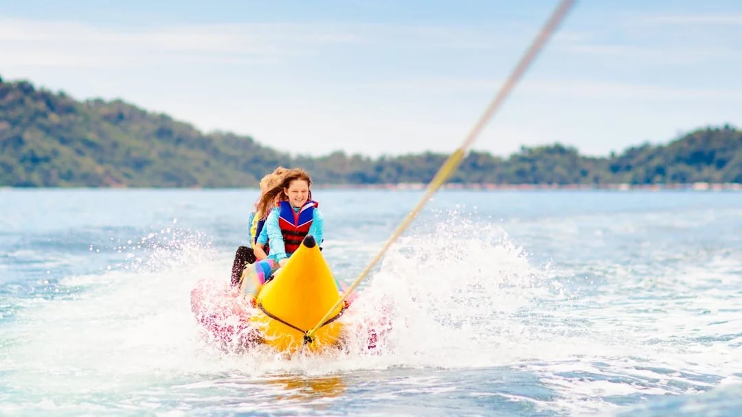 Kids enjoying a banana boat ride on the ocean with distant hills in the background.