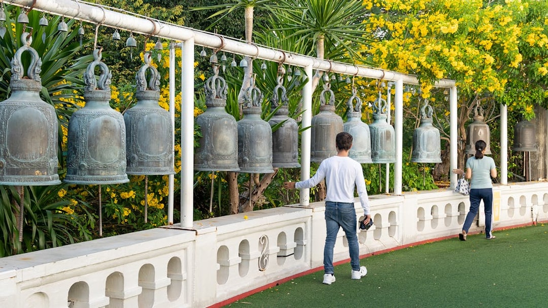 Visitors ringing bells at Wat Pho, Bangkok, surrounded by lush greenery and yellow flowers.