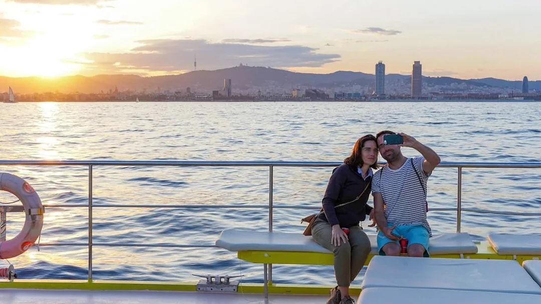 Couple taking a selfie on a catamaran cruise with Barcelona skyline at sunset.