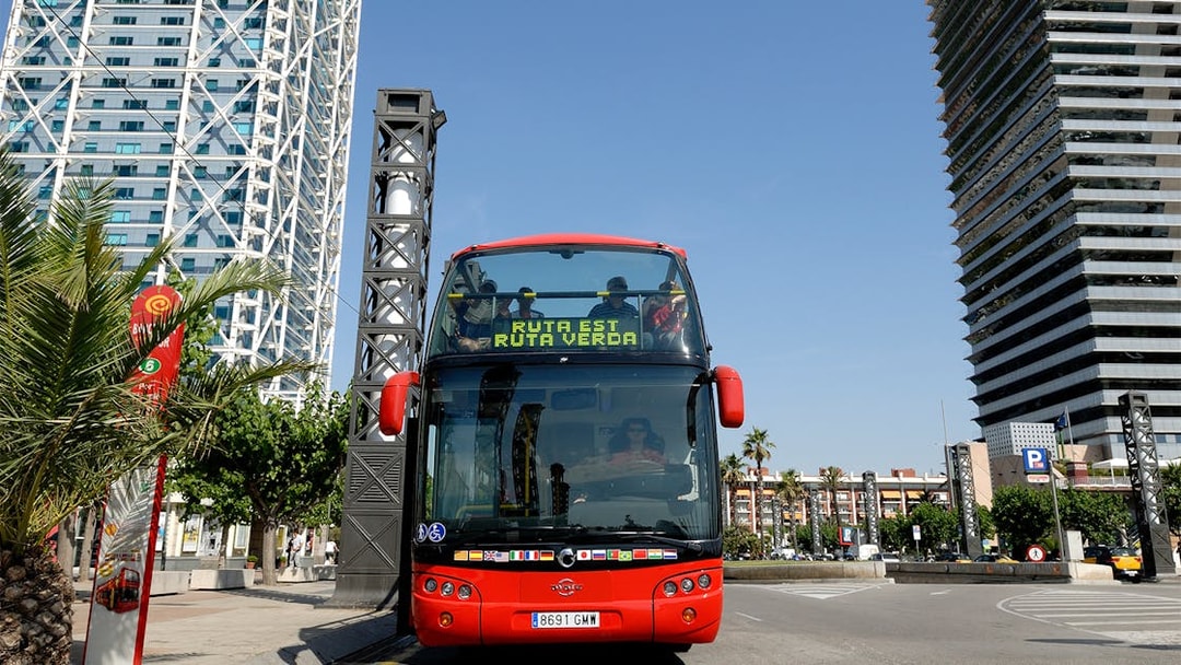 Red double-decker bus on Barcelona City Hop On Hop Off Tour near modern buildings.