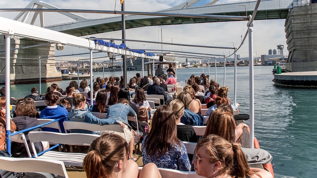 Passengers on Las Golondrinas boat trip under a bridge in Barcelona harbor.