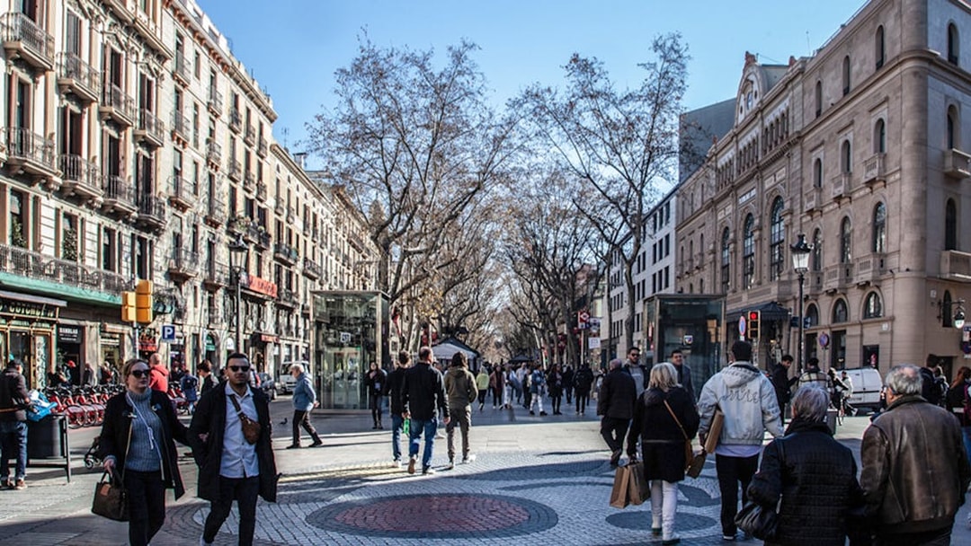 People walking along Las Ramblas in Barcelona, with historic buildings and trees lining the street.