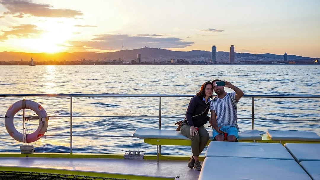 Couple taking selfie on catamaran cruise with Barcelona skyline at sunset.