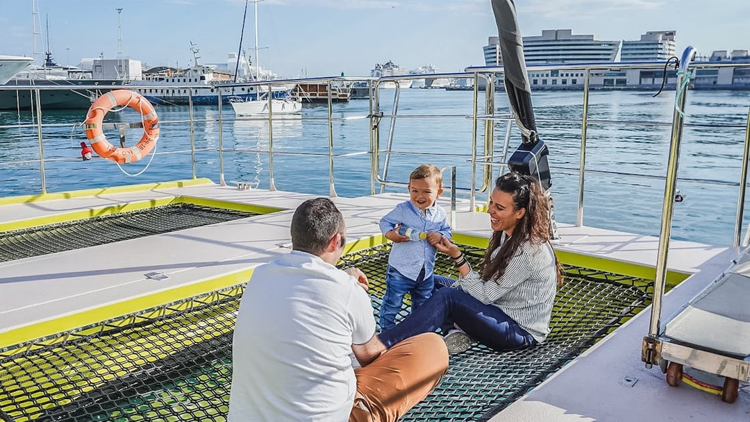 Tourists relaxing on catamaran cruise deck with child, harbor view in background.