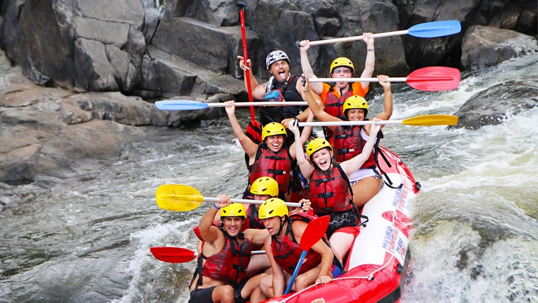 Group rafting on Barron River, navigating rapids with paddles raised, wearing helmets and life vests.