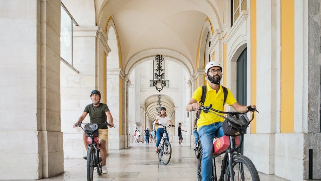 Cyclists riding e-bikes through an arched corridor in Lisbon, Portugal.