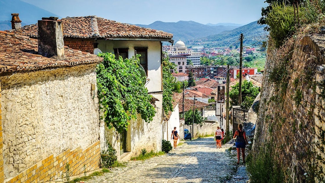 Guests walking along cobblestone street in Berat Old Town, Albania, with historic buildings and distant hills.