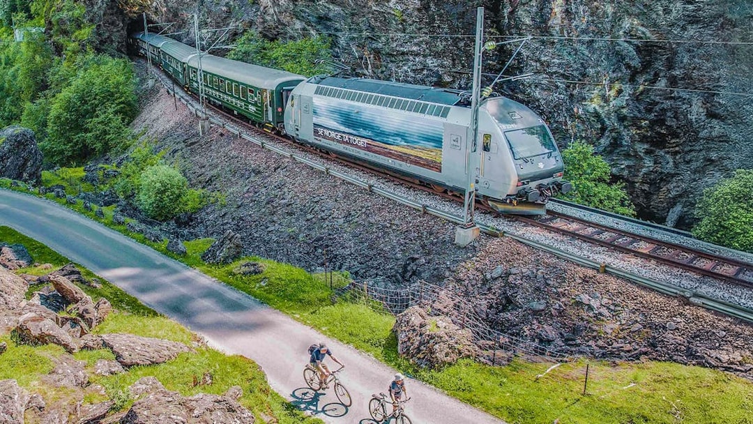 Flåm Railway train passing through rocky landscape with cyclists on nearby path.