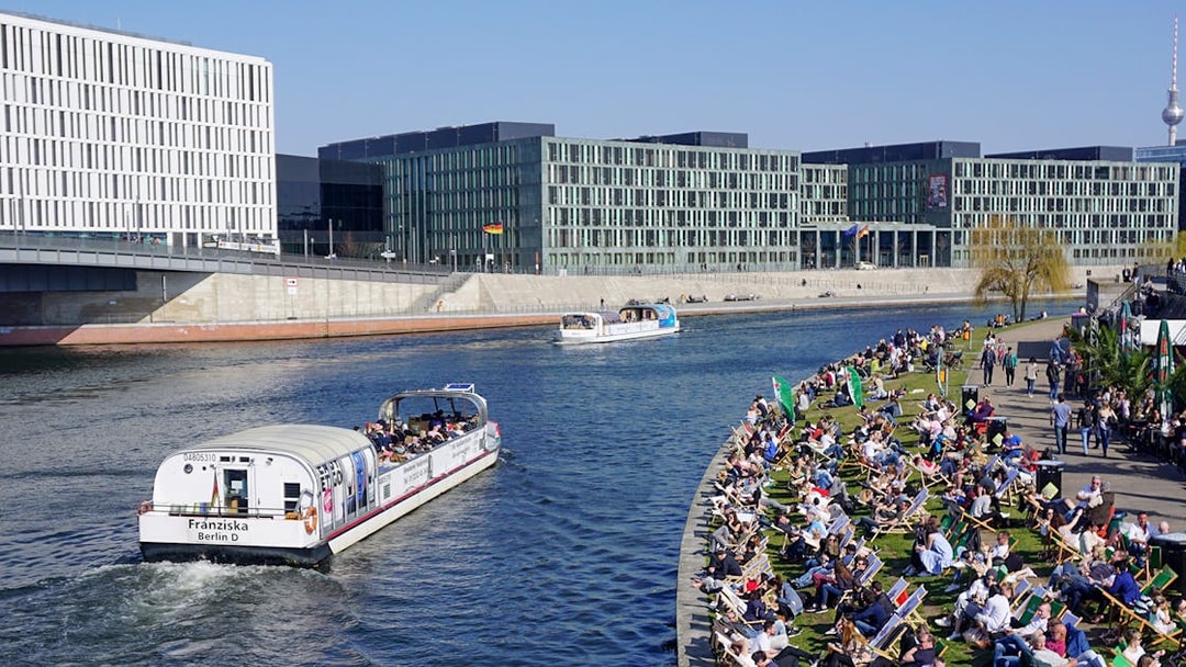 River cruise boat on Spree River in Berlin with passengers and riverside crowd.