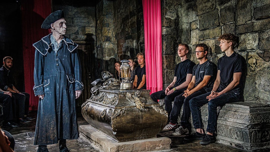Guide in historical costume leading a tour inside Berlin Dungeon with visitors seated around a tomb.