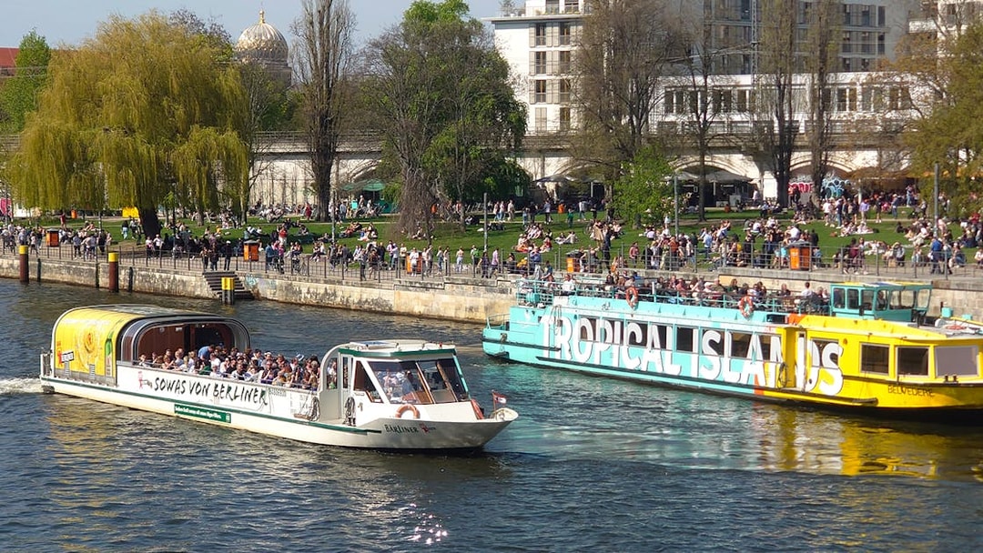 Sightseeing cruise boats on the Spree River in Berlin with people enjoying the riverside.