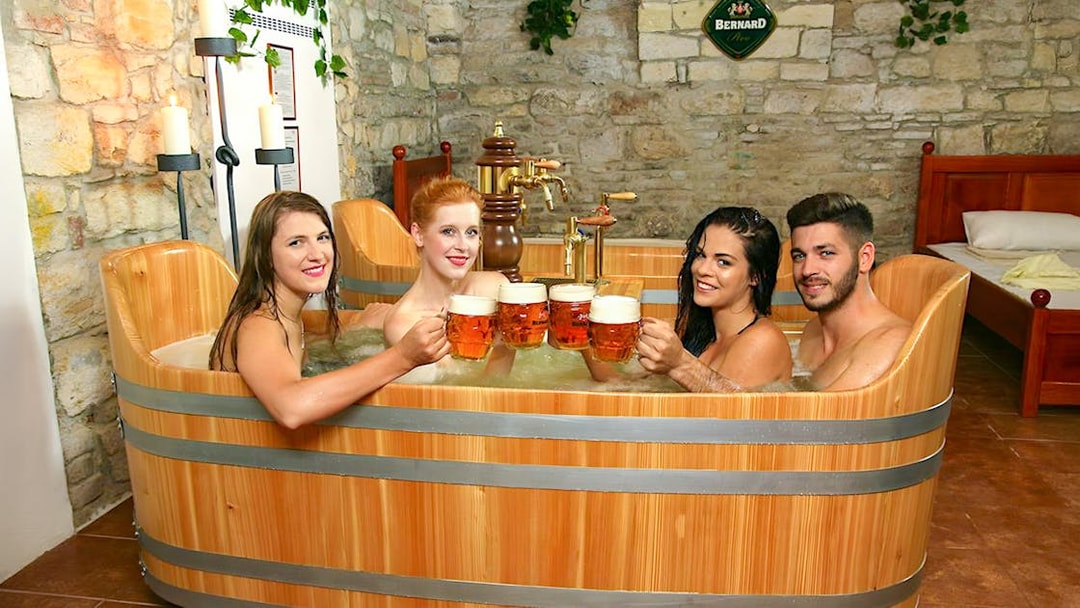 Guests enjoying a beer bath at Bernard Beer Spa, raising mugs in a wooden tub.