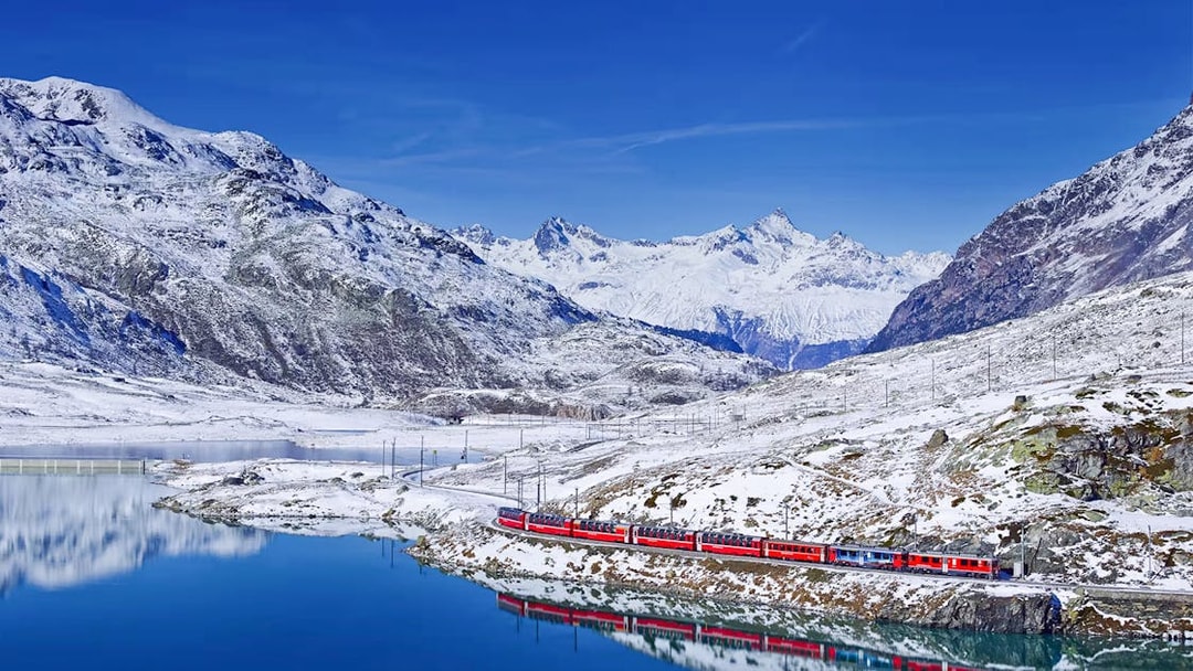 Bernina Express train traveling through snowy Swiss Alps with mountain reflection in lake.