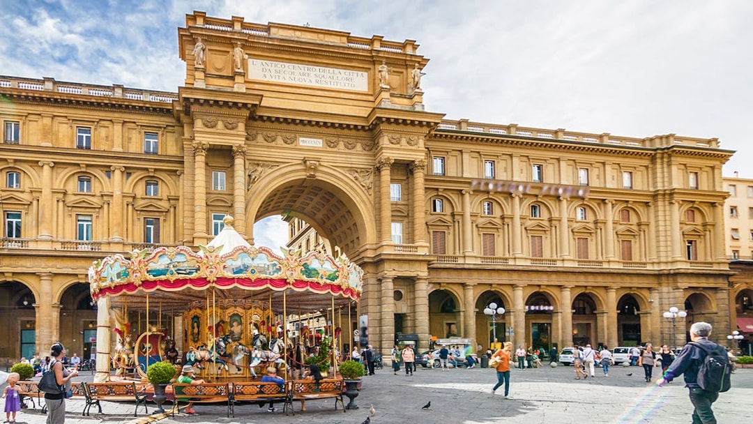 Carousel in Republic Square, Florence, with historic arch and people walking nearby.