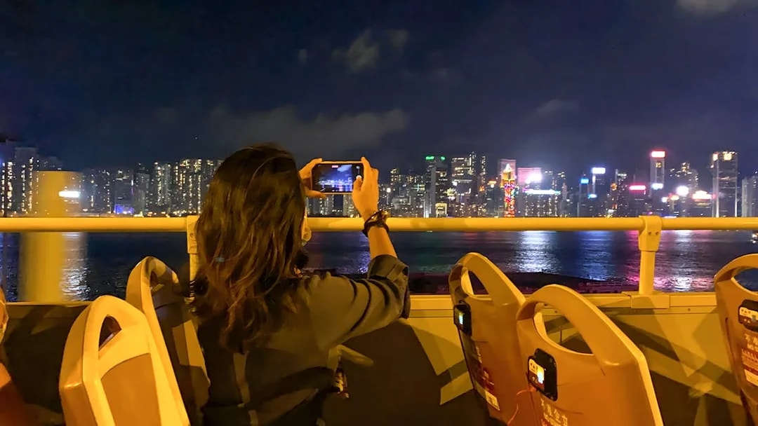 Person taking photo of Hong Kong skyline from open-top bus at night.