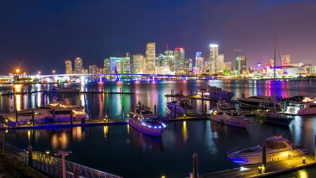 Miami skyline at night with illuminated buildings and yachts in the foreground.