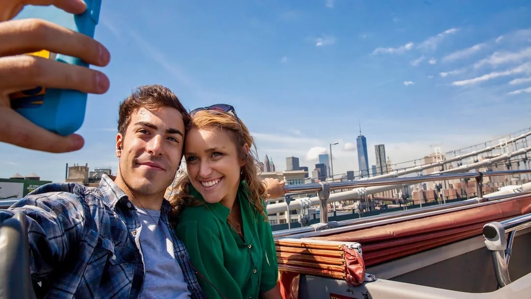 Couple taking selfie on open-top bus with New York skyline in background.