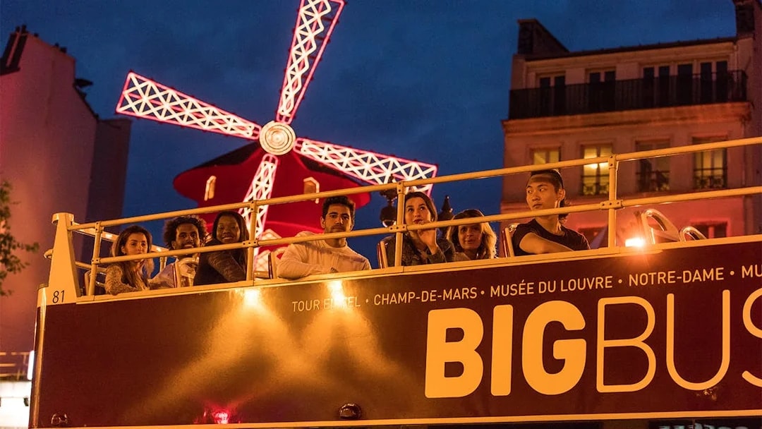 Open-top bus tour passing Moulin Rouge at night in Paris.