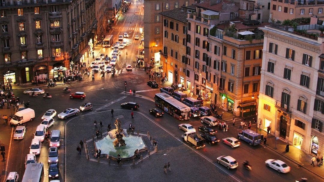 Rome cityscape at night with busy streets and illuminated buildings, featuring a central fountain.