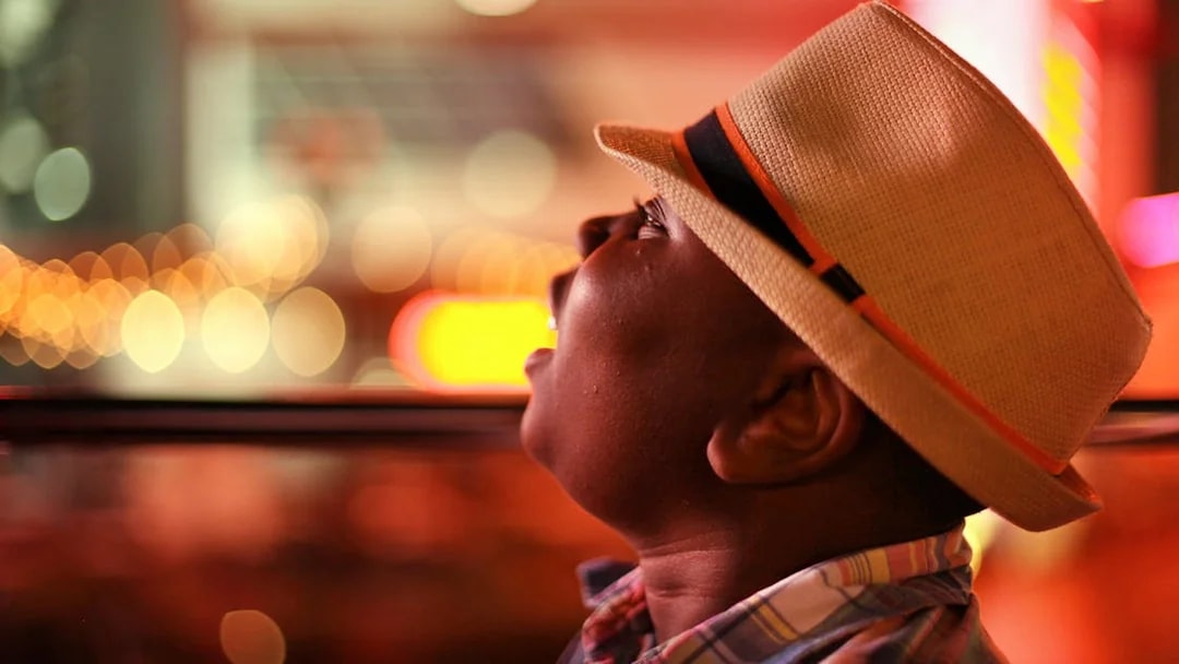 Child wearing a hat looking out of a bus window at night city lights.