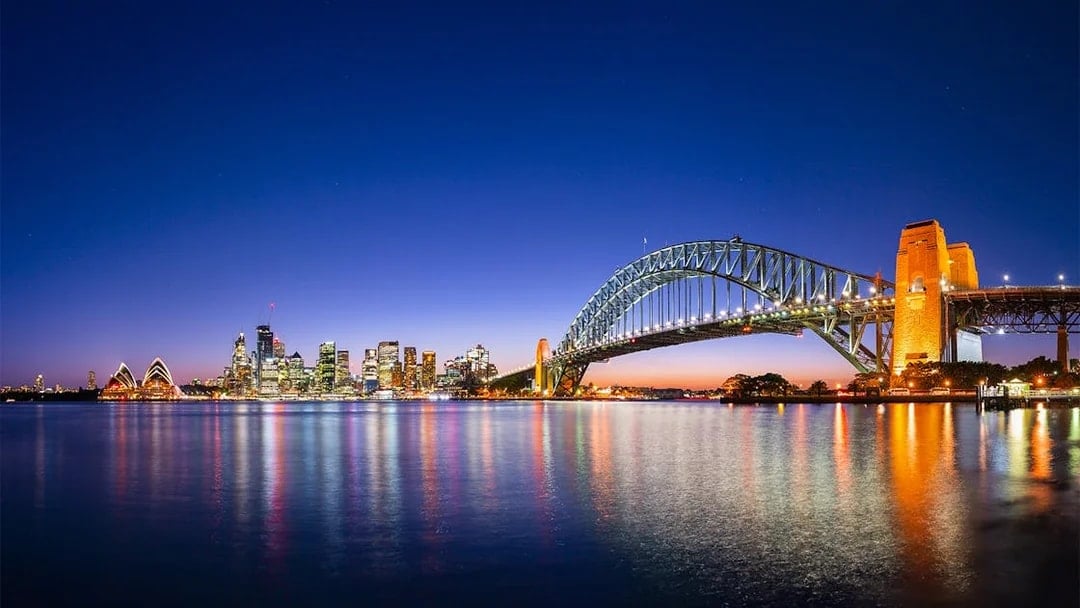 Sydney Harbour Bridge and Opera House illuminated at night with city skyline reflected in the water.