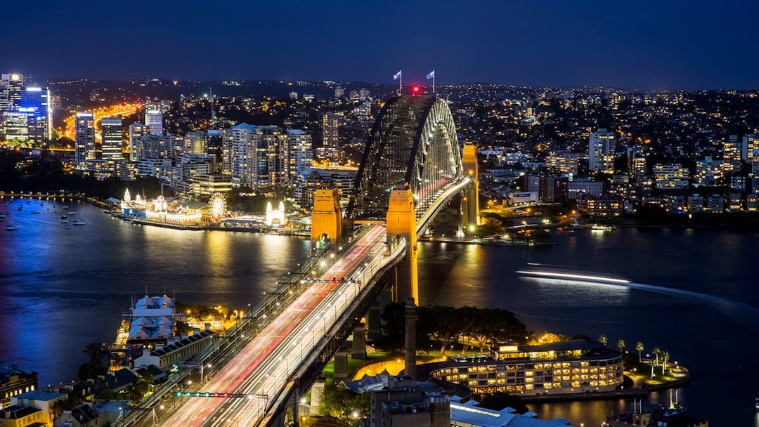 Sydney Harbour Bridge illuminated at night with city skyline and water reflections.