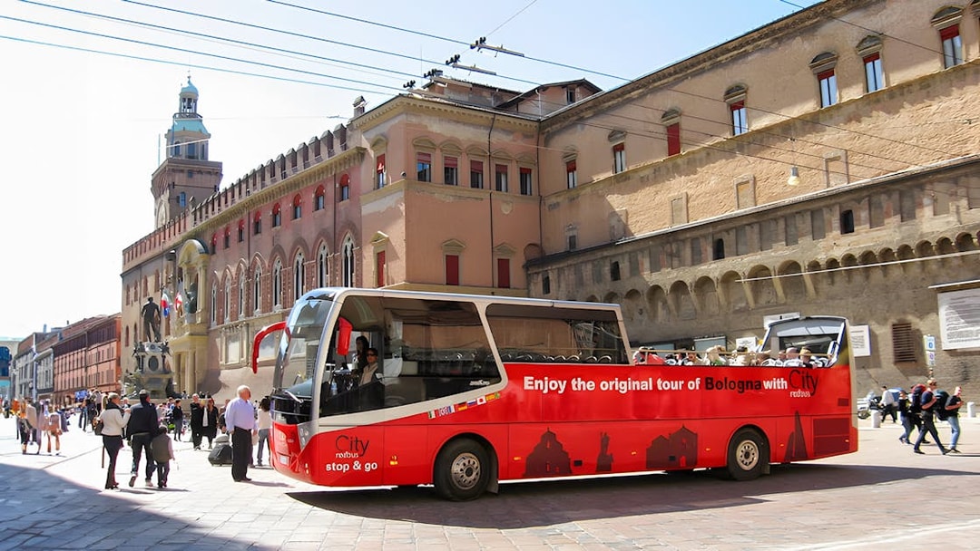 Red city tour bus in Bologna near historic buildings.