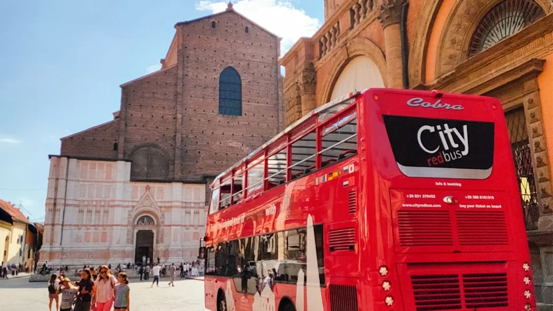 Red double-decker bus near historic church in Bologna, Italy, with tourists nearby.