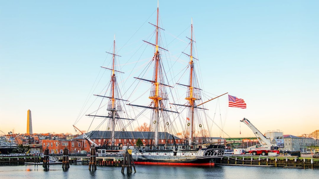 USS Constitution docked in Boston with American flag and cityscape in the background.