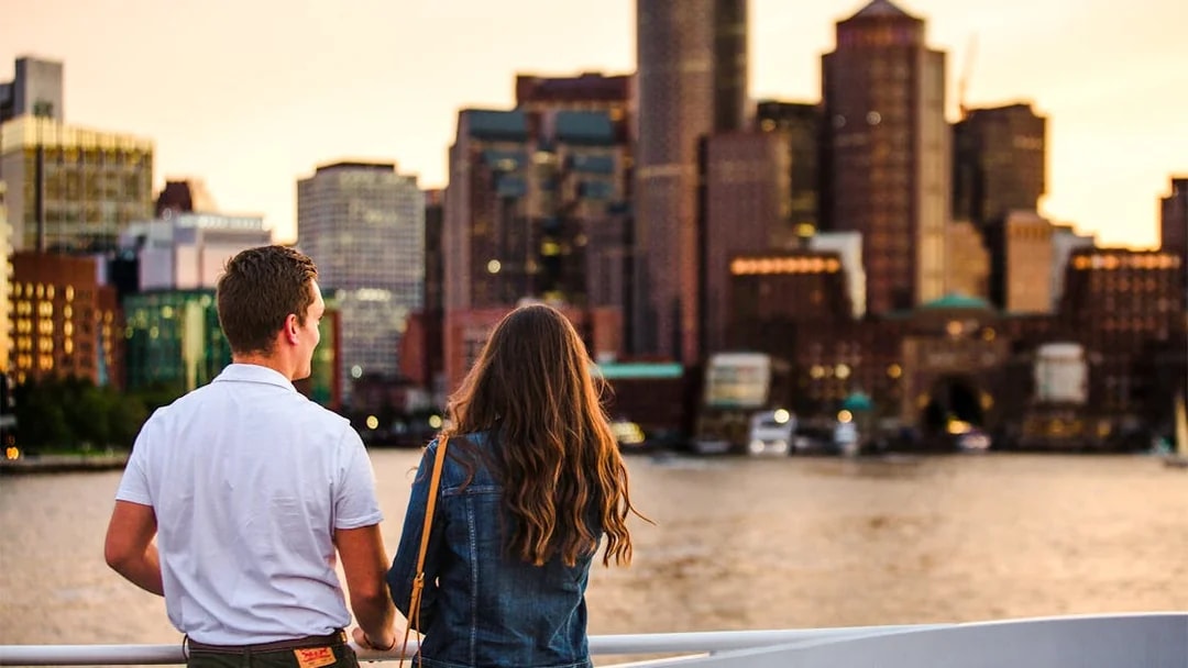 Guests enjoying a Boston sunset cruise with city skyline in the background.