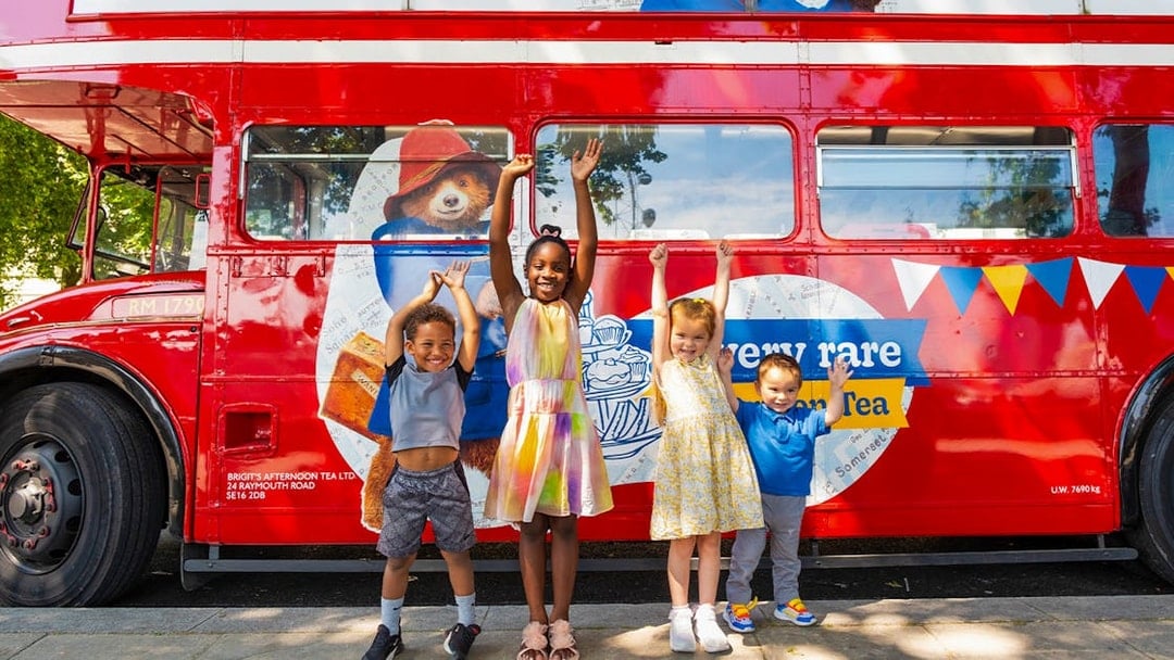 Children smiling in front of the Paddington Afternoon Tea Bus in London.