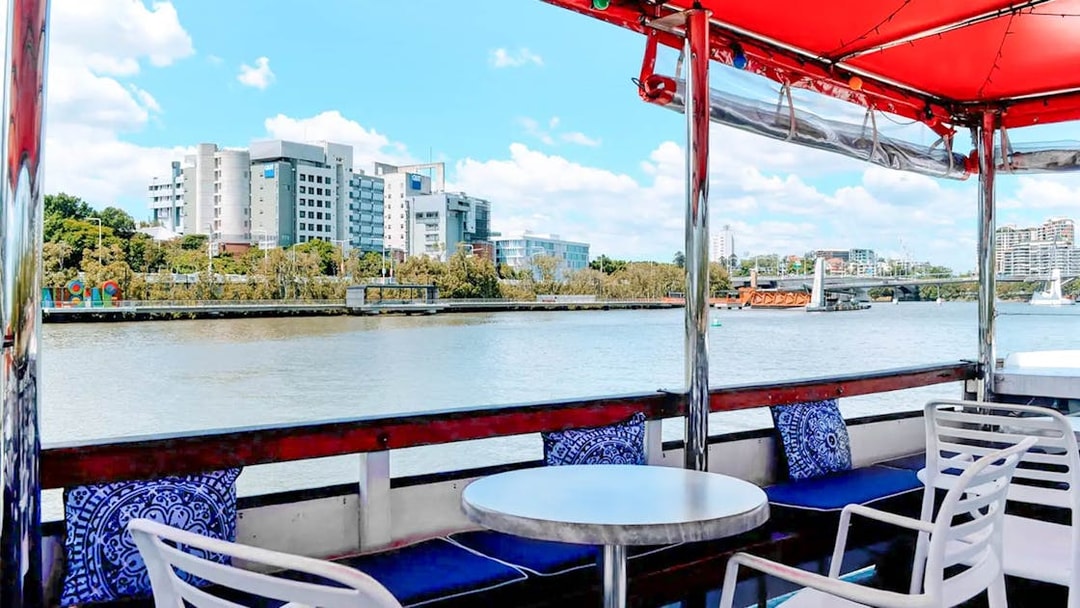 Covered deck view of Brisbane River cruise with city buildings and bridge in the background.