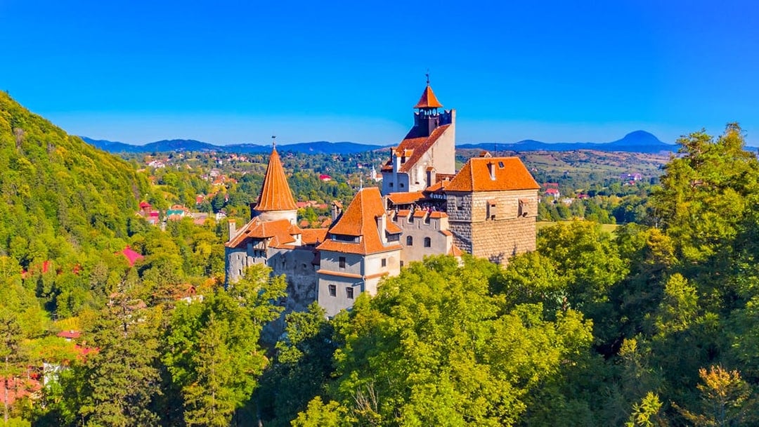 Bran Castle in Romania surrounded by lush greenery and distant hills.