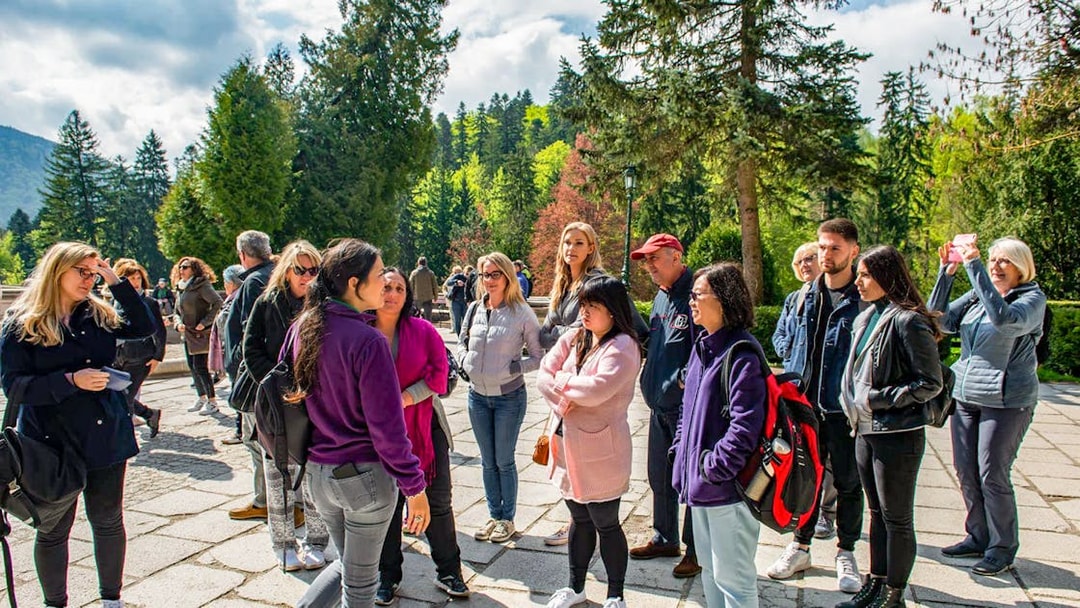 Group of tourists listening to a guide at Bran Castle, surrounded by lush greenery.