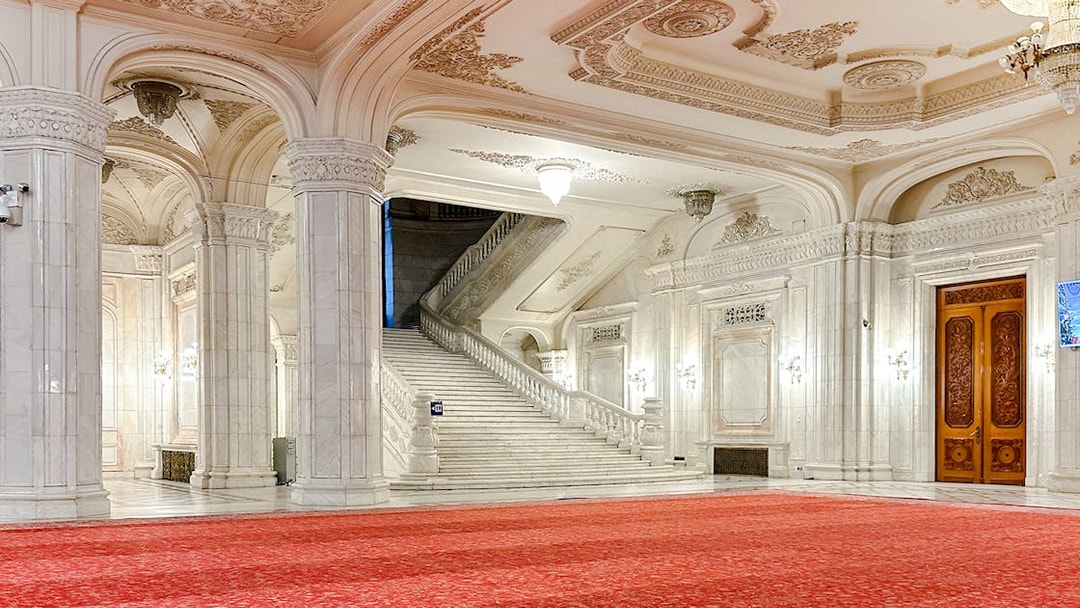 Grand staircase in the Palace of Parliament, Bucharest, with ornate ceilings and red carpet.