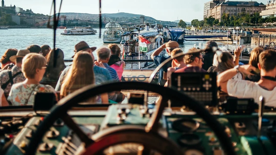 Danube River cruise with tourists seated, Budapest skyline in background.